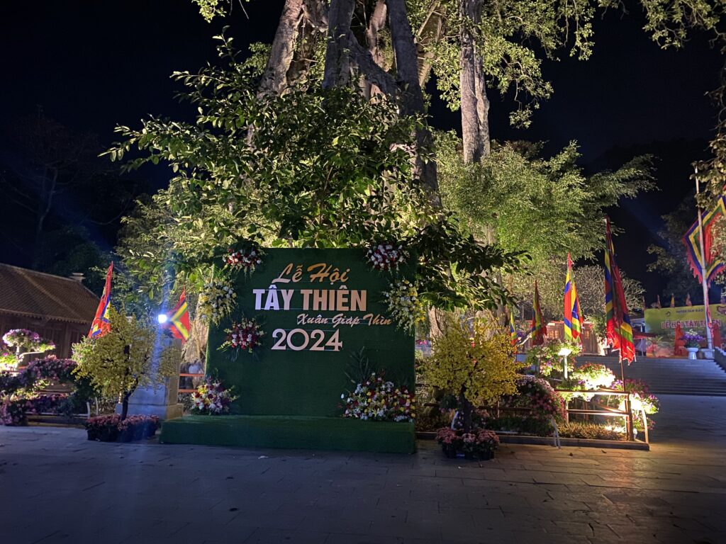 Tiểu cảnh Lễ hội Tây Thiên 2024 với phông xanh, hoa tươi và cây mai trang trí vào buổi tối.
